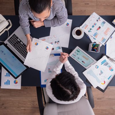 Top view of office workers meeting analyzing financial charts holding clipboard. Manager and employer discussing marketing strategy during briefing. Pointing at graphs.